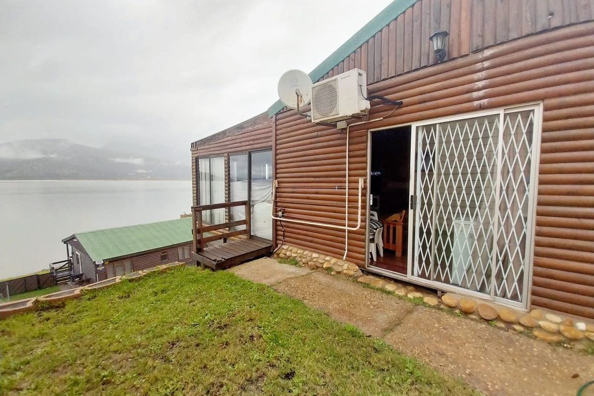 Rear exterior view of a log cabin featuring sliding glass doors with security gates, an air conditioning unit, and scenic waterfront views.