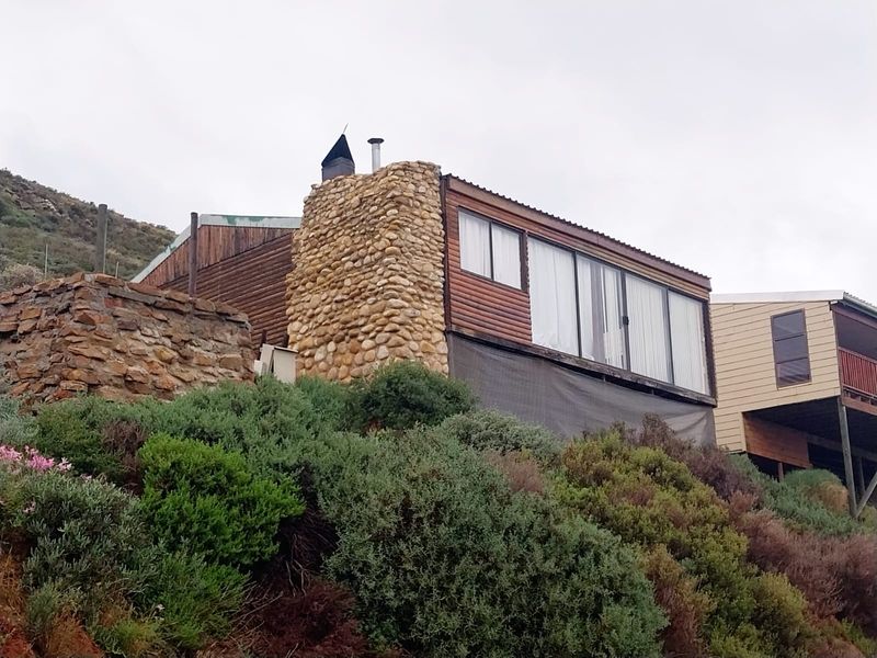 Exterior view of a hillside property featuring a prominent stone chimney, timber cladding, and elevated sliding glass doors overlooking the landscape.
