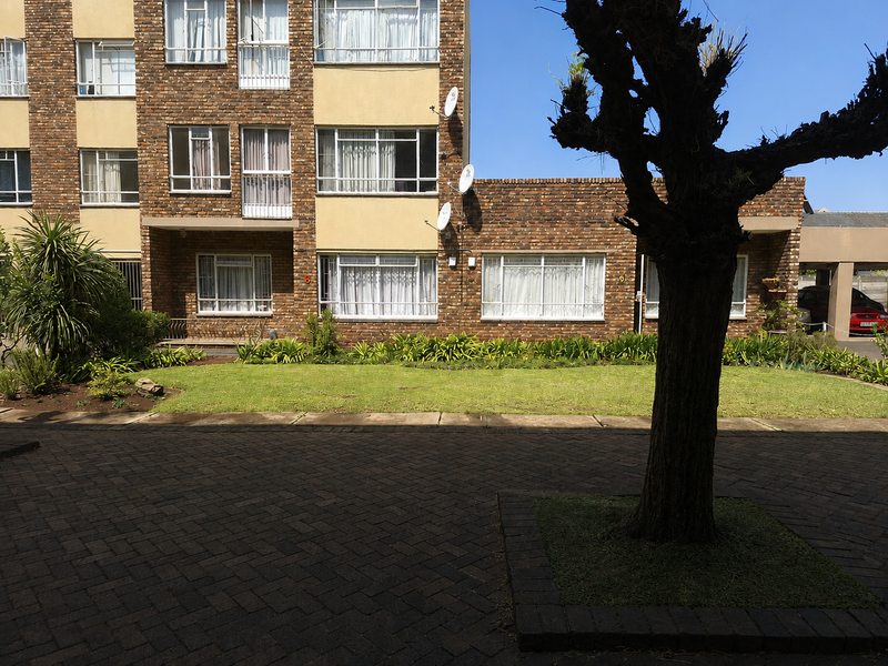 A view of the apartment building exterior featuring a brick facade, paved courtyard area, and a well-maintained garden space.