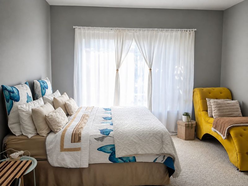 A well-maintained bedroom featuring neutral grey walls, carpeted flooring, and a large window dressed with sheer curtains.