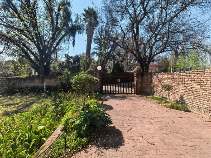 A front entrance view featuring a paved driveway, mature trees, landscaped garden beds, and a secure metal gate set within a brick boundary wall.