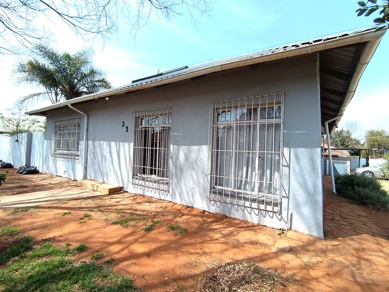 Front elevation of a single-story house featuring security bars on windows, a metal roof with solar water heating, and stone paved ground area.