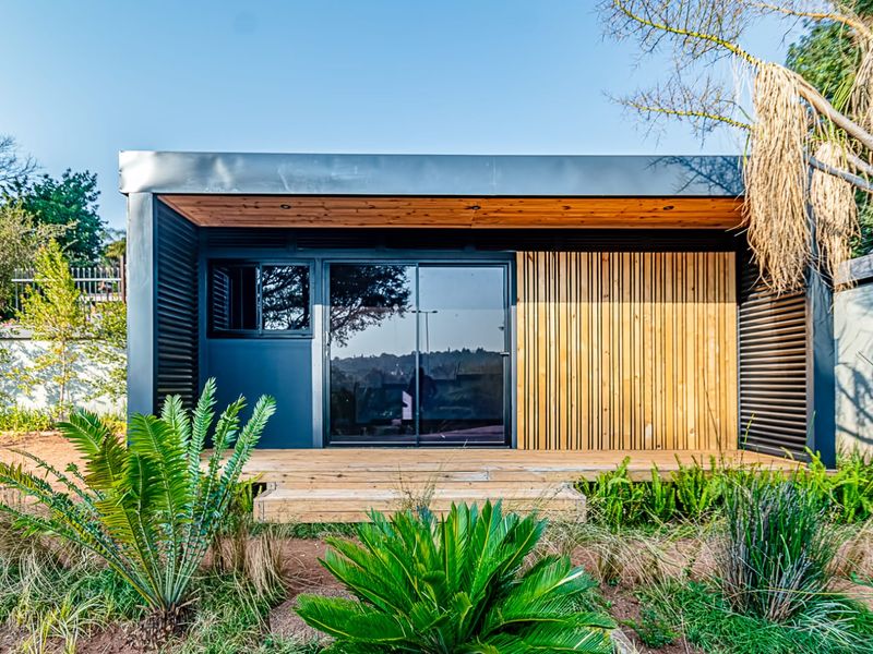 A modern, compact residential structure featuring timber decking, vertical wood slat cladding, and a sleek metal roof, set within a landscaped garden.