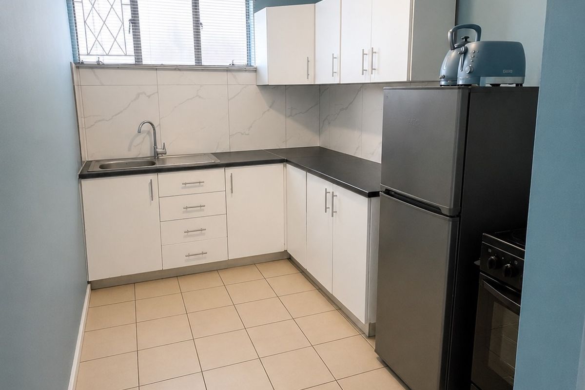 A functional kitchen featuring white cabinetry, dark countertops, a stainless steel sink, tiled backsplash, and ceramic floor tiles with security bars on the window.