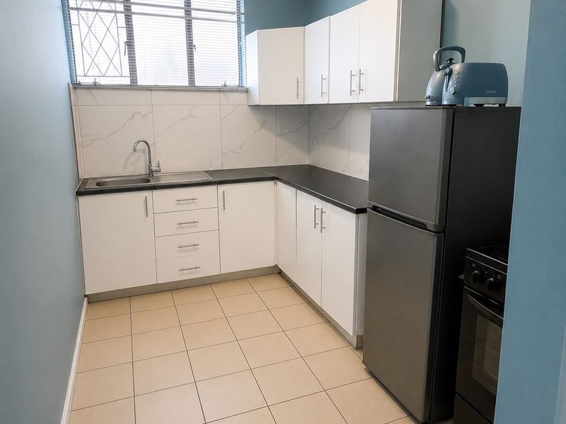 A functional kitchen featuring white cabinetry, dark countertops, a stainless steel sink, tiled backsplash, and ceramic floor tiles with security bars on the window.