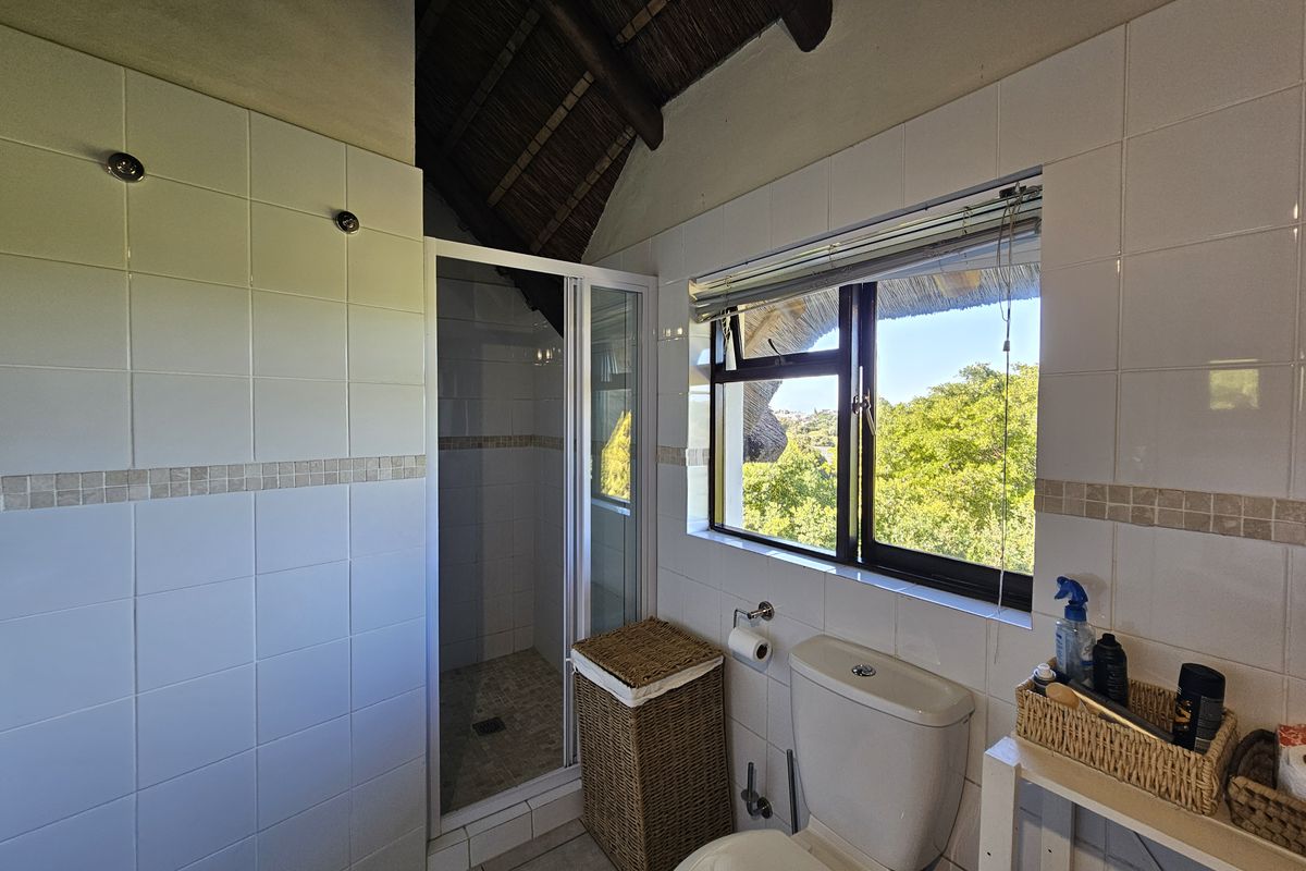 A bathroom featuring a shower enclosure, toilet, and tiled finishes, set under an exposed thatch roof with a window overlooking greenery.