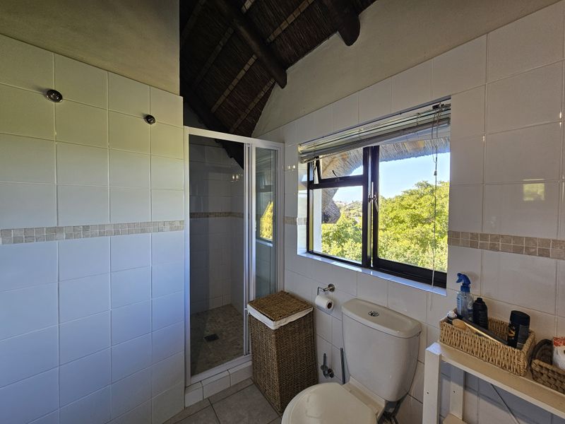A bathroom featuring a shower enclosure, toilet, and tiled finishes, set under an exposed thatch roof with a window overlooking greenery.
