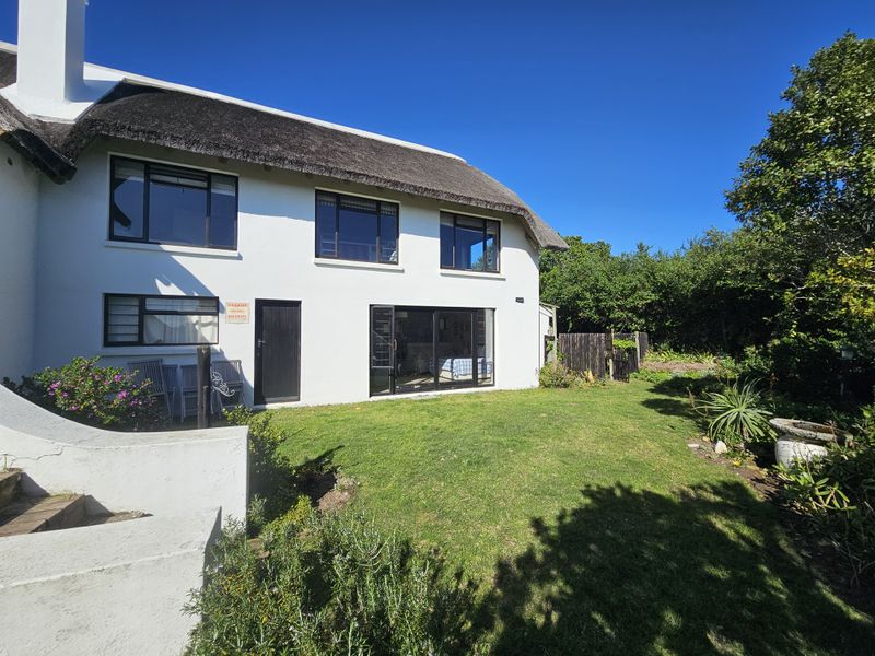 Rear view of a white-walled house featuring a traditional thatched roof, large sliding glass doors, and a well-maintained garden lawn.