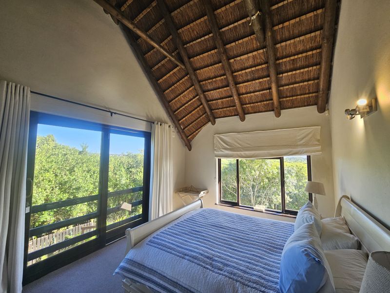 A bright bedroom featuring a high vaulted thatched ceiling with exposed timber rafters, large windows, and sliding glass doors offering scenic views.