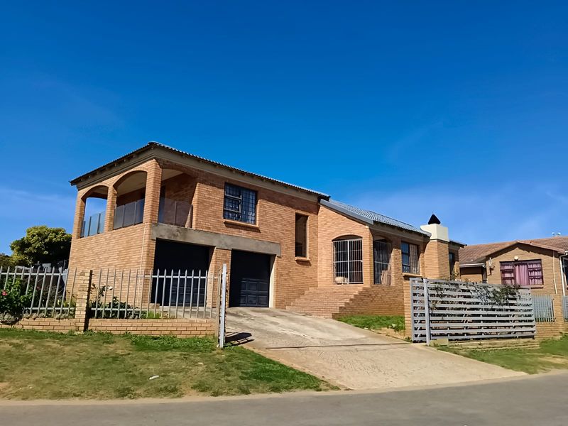 A double-story face brick house featuring a double garage, an upper-level balcony with glass balustrades, and a paved driveway behind secure metal fencing.