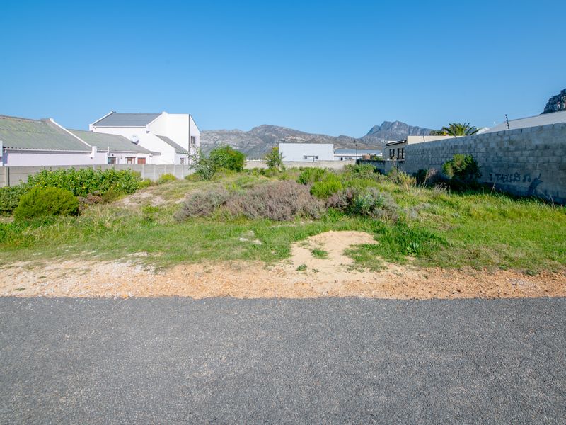 A vacant residential plot featuring a level surface, boundary walls, and mountain views, situated along an established asphalt road.