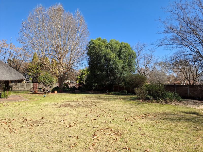 A spacious backyard garden featuring a large lawn, mature trees, a perimeter wall, and a corner thatched lapa area.