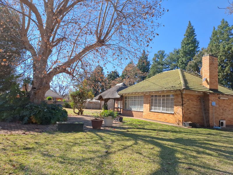Rear view of a brick house featuring a tiled roof, chimney, and a paved patio area adjacent to a garden with a thatched lapa.