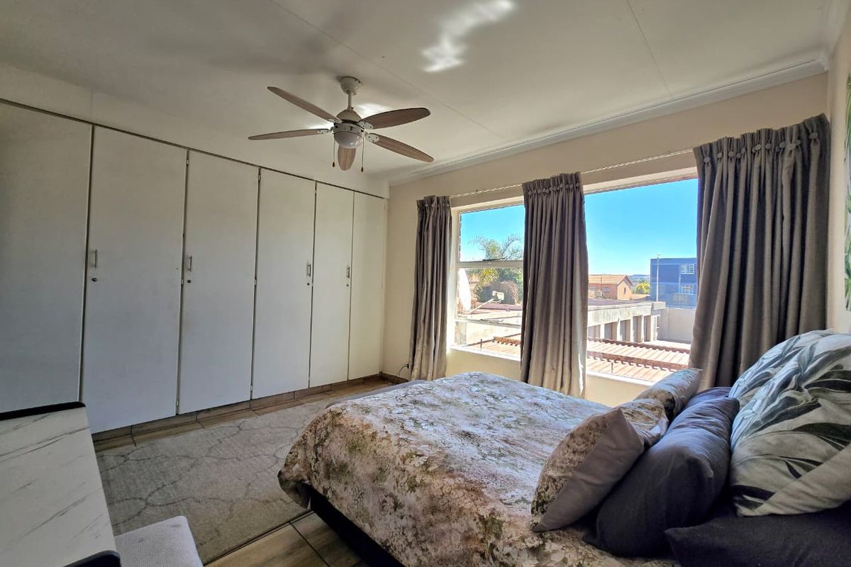 A well-lit bedroom featuring extensive built-in cupboards, a ceiling fan, and large windows providing natural light and an urban view.