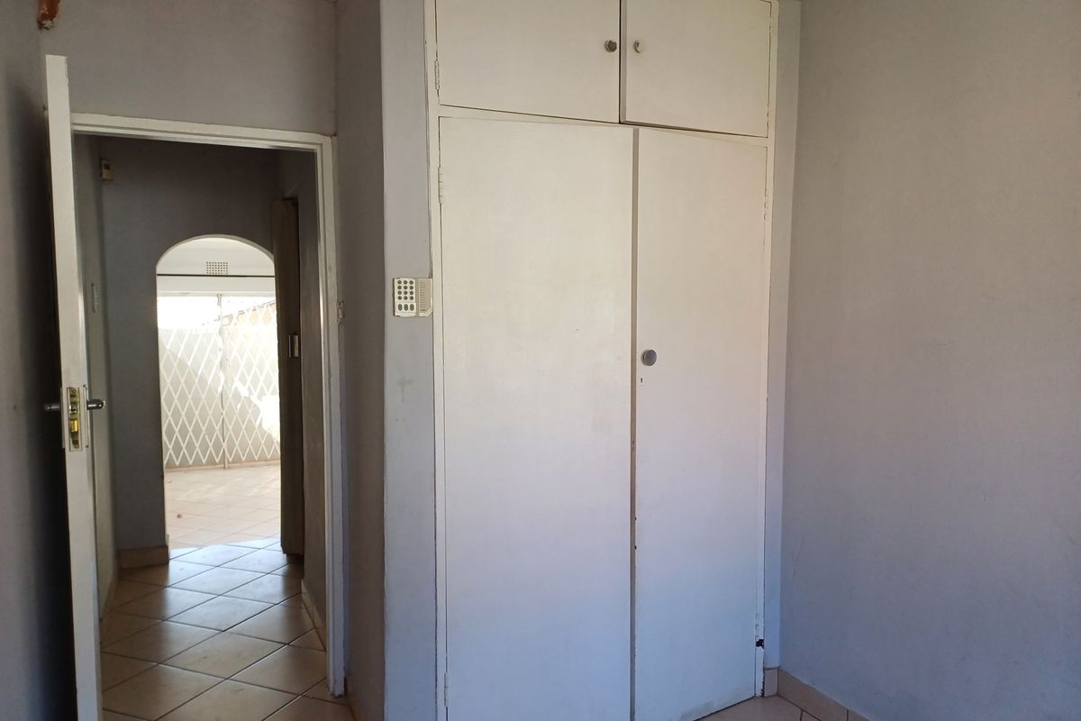 An empty bedroom featuring built-in cupboards, tiled flooring, and an alarm keypad, with an adjacent hallway leading to an outdoor area.