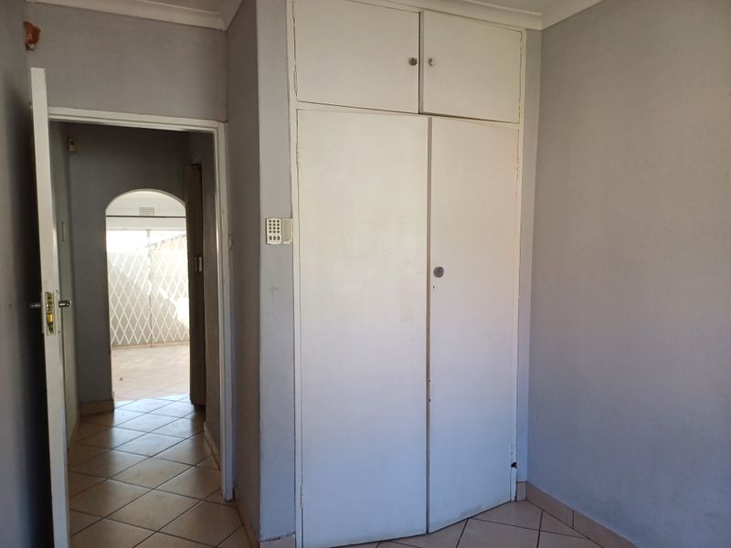 An empty bedroom featuring built-in cupboards, tiled flooring, and an alarm keypad, with an adjacent hallway leading to an outdoor area.