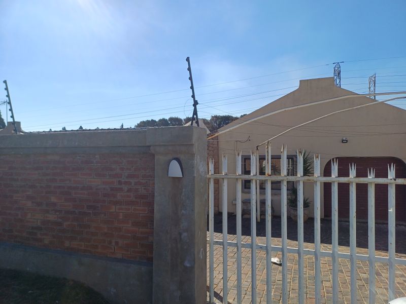 Front exterior view featuring a brick boundary wall with electric fencing, a metal security gate, and a paved driveway leading to the garage.