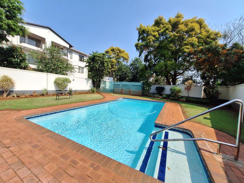 A well-maintained outdoor swimming pool area featuring brick paving, a metal handrail, and surrounding landscaped garden space within a secure complex.