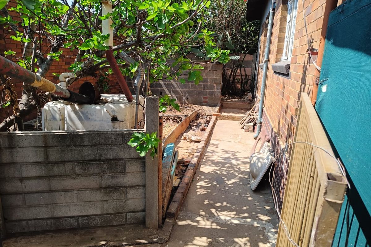A narrow rear exterior side passage featuring brick and cinder block walls, a concrete pathway, and a damaged water tank.