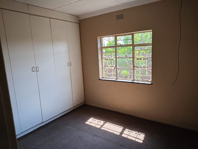 A vacant bedroom featuring built-in cupboards, carpeted flooring, and a steel-framed window with security bars.