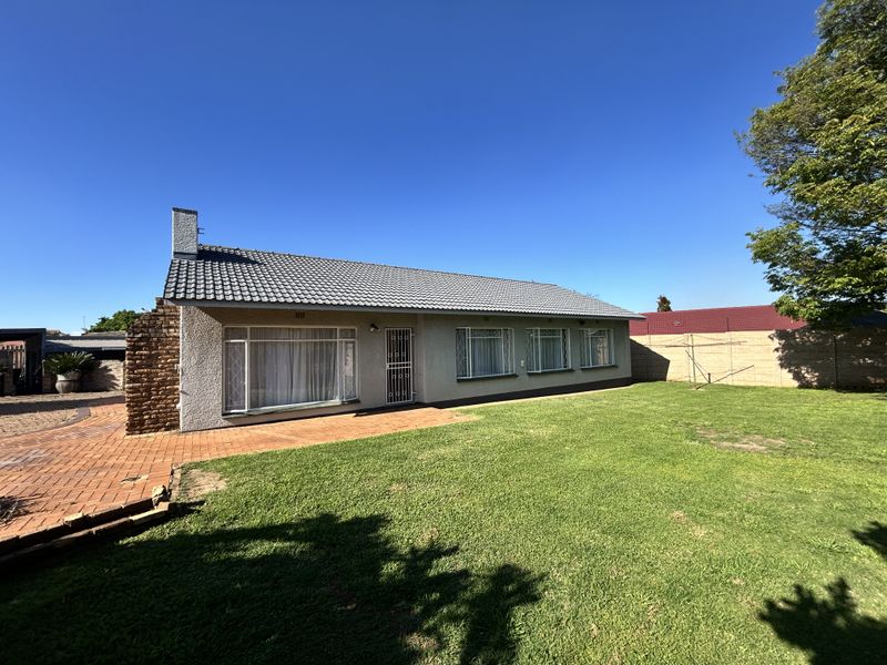Rear view of a single-story house featuring a tiled roof, brick chimney, paved patio area, and a spacious lawn with perimeter walling.