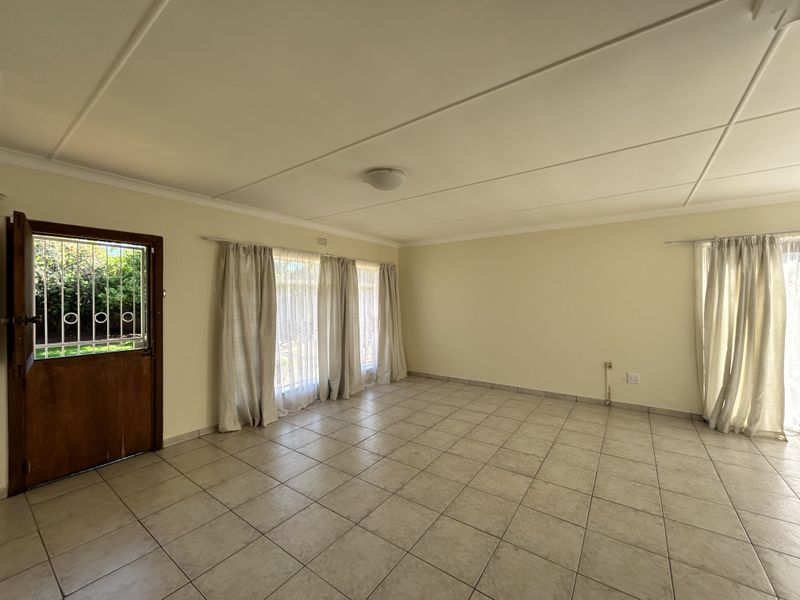 A spacious, empty living room featuring neutral tiled flooring, large windows with curtains, and a secure timber entrance door.