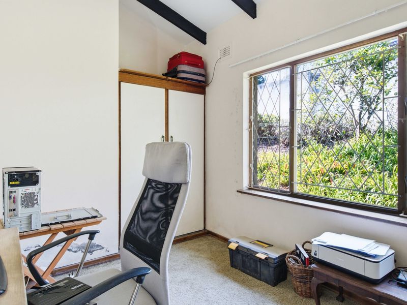 A home office featuring built-in cupboards, exposed ceiling beams, and a large leaded glass window overlooking the garden.