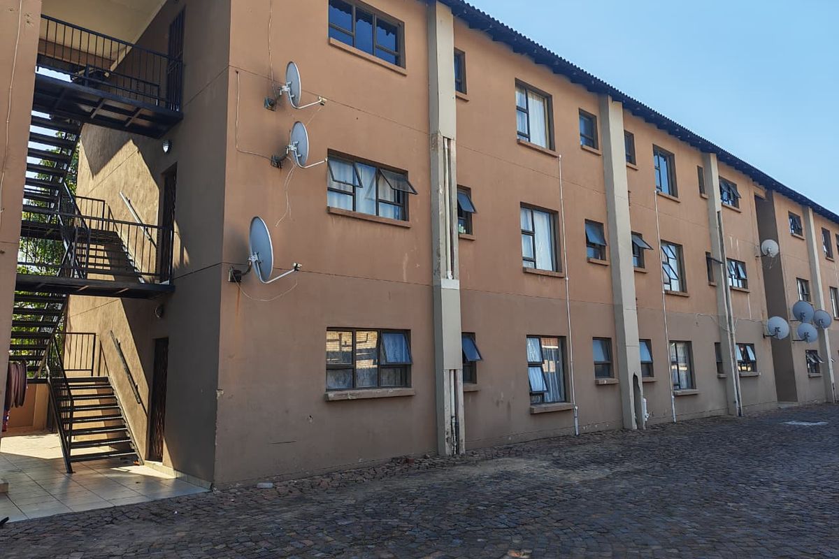 Exterior view of a multi-story apartment block featuring a metal staircase, paved courtyard area, and multiple satellite dish installations on the facade.