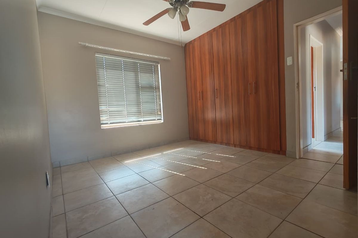 A spacious, empty bedroom featuring tiled flooring, built-in wooden cupboards, a ceiling fan, and natural light through venetian blinds.
