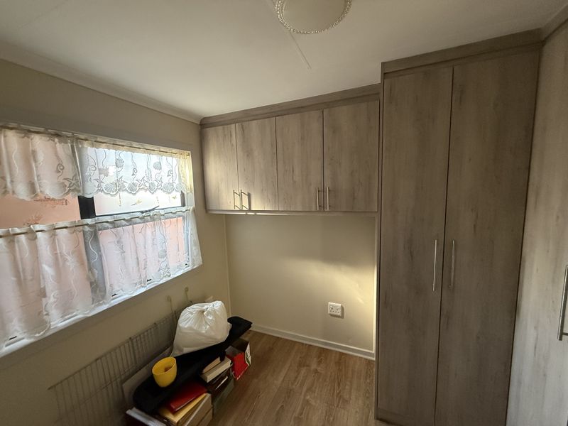 A bedroom featuring light wood-finish built-in cupboards and overhead storage units, complemented by laminate flooring and a window with natural light.