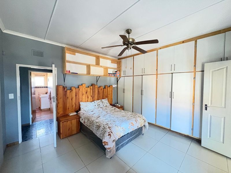 Spacious bedroom featuring extensive built-in cupboards, overhead shelving, a ceiling fan, and tiled flooring, with an adjoining en-suite bathroom.