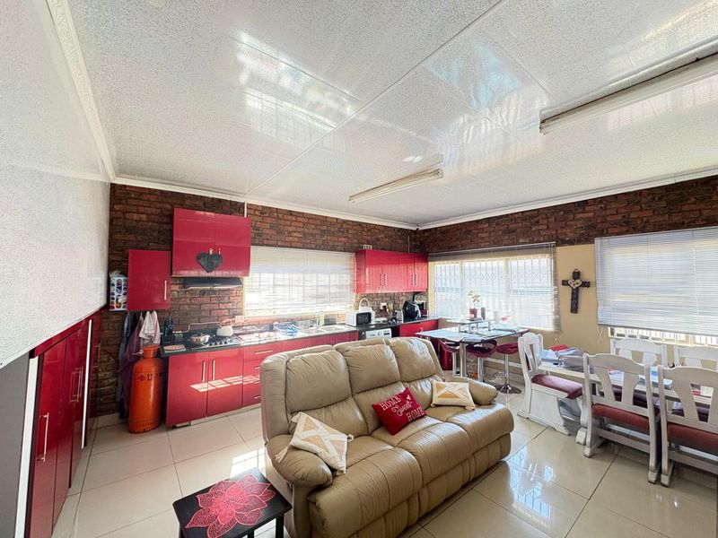 Open-plan kitchen and living area featuring red cabinetry, exposed brick walls, tiled flooring, and large windows with security bars.