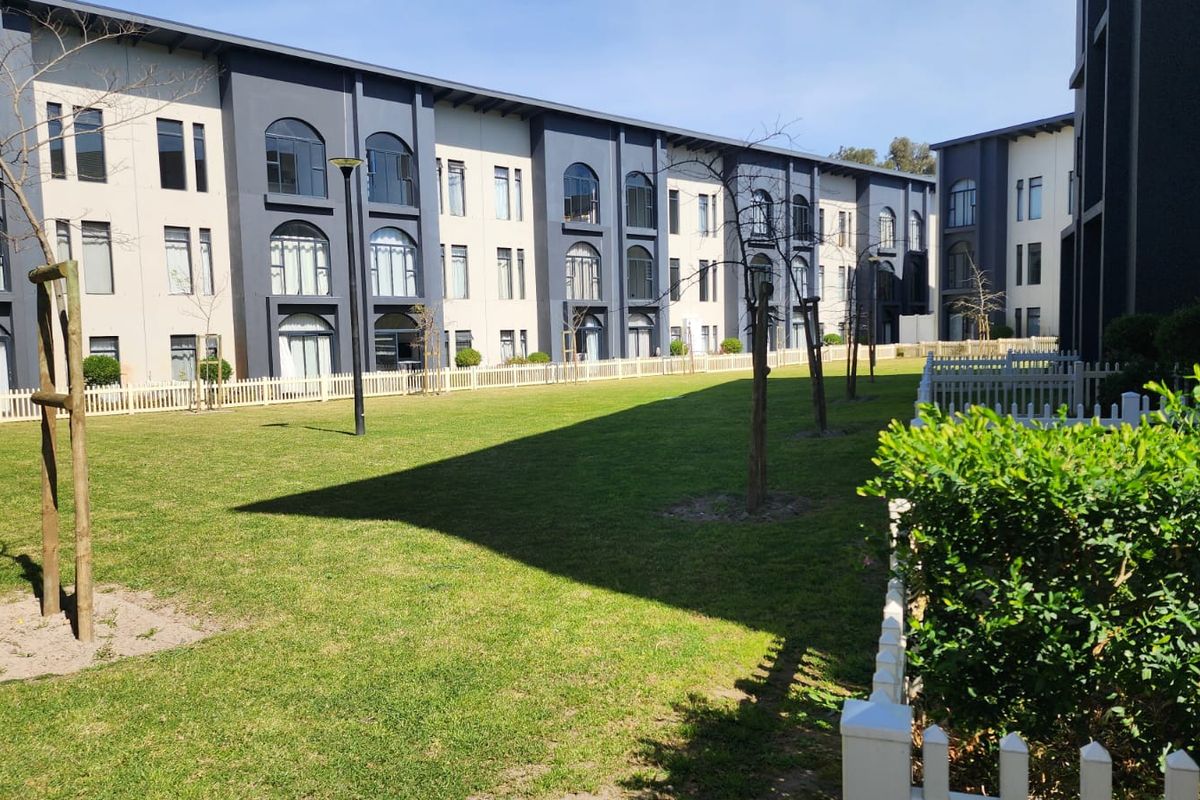 A well-maintained communal garden area featuring manicured lawns, picket fencing, and a modern multi-story apartment building facade with arched window detailing.
