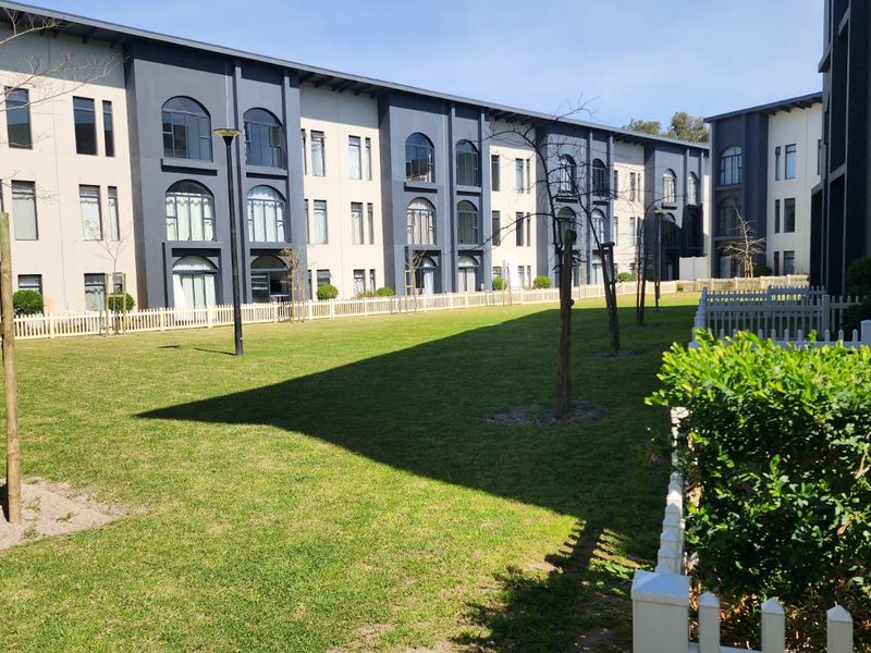 A well-maintained communal garden area featuring manicured lawns, picket fencing, and a modern multi-story apartment building facade with arched window detailing.
