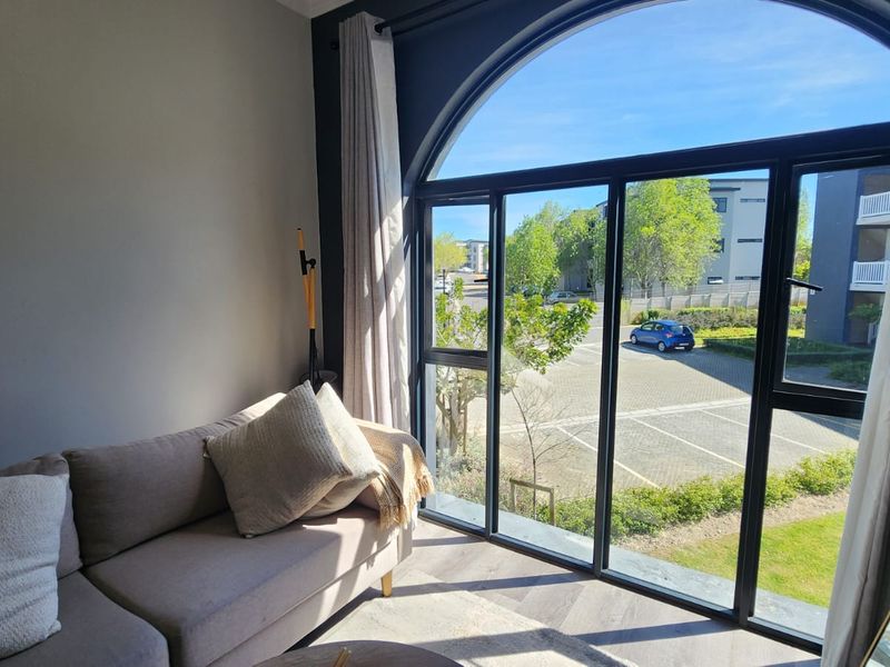 A bright living area featuring a large arched window with black aluminium frames and modern herringbone flooring, offering a pleasant view of the complex.