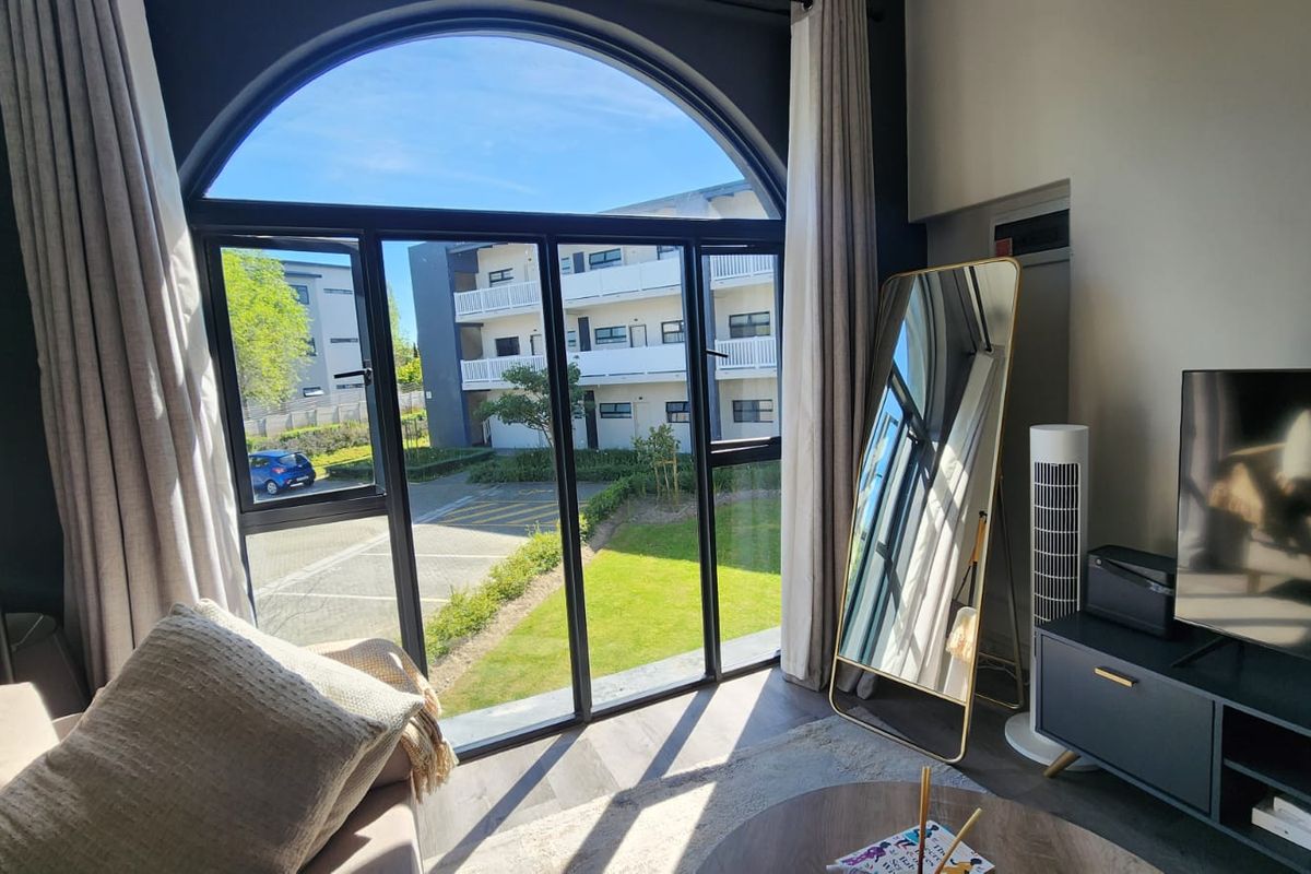 A bright living room featuring a large arched window with black aluminium frames, offering views of the complex grounds and laminate flooring.