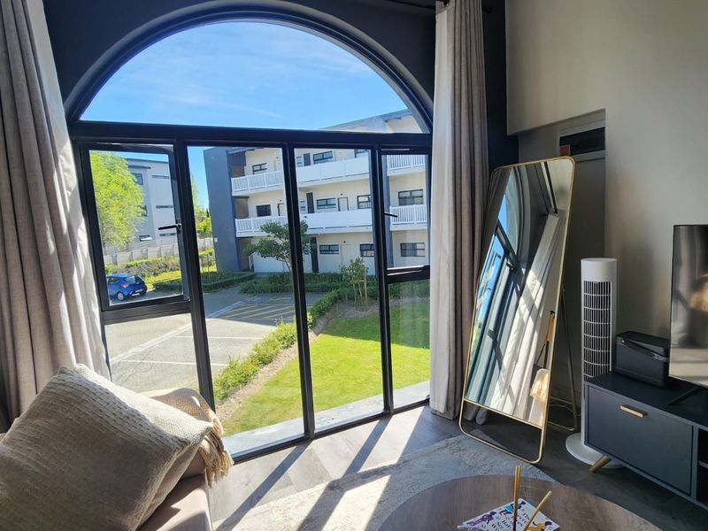 A bright living room featuring a large arched window with black aluminium frames, offering views of the complex grounds and laminate flooring.