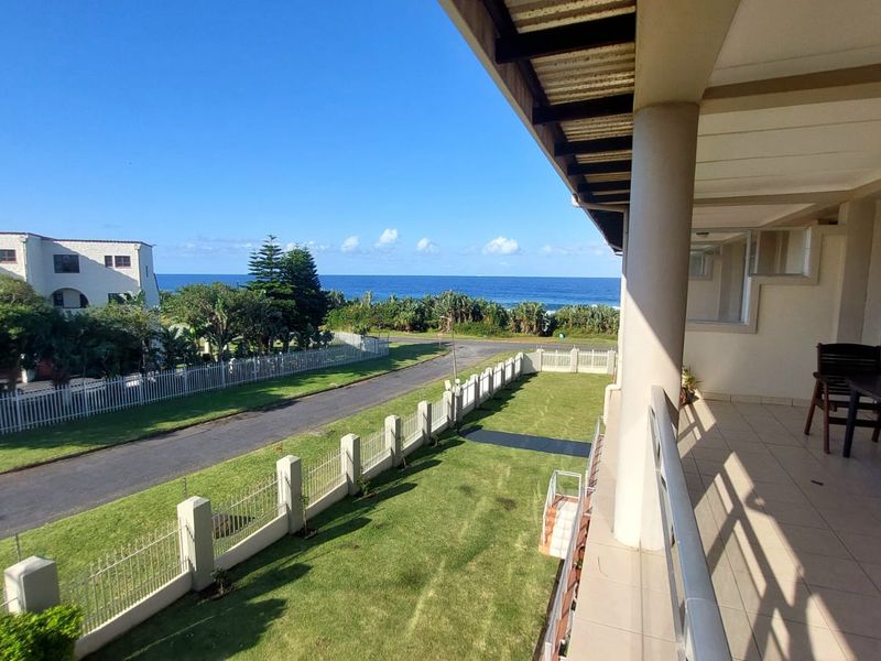 A spacious covered balcony featuring tiled flooring and metal railings, offering an elevated view of the surrounding grounds and the ocean.