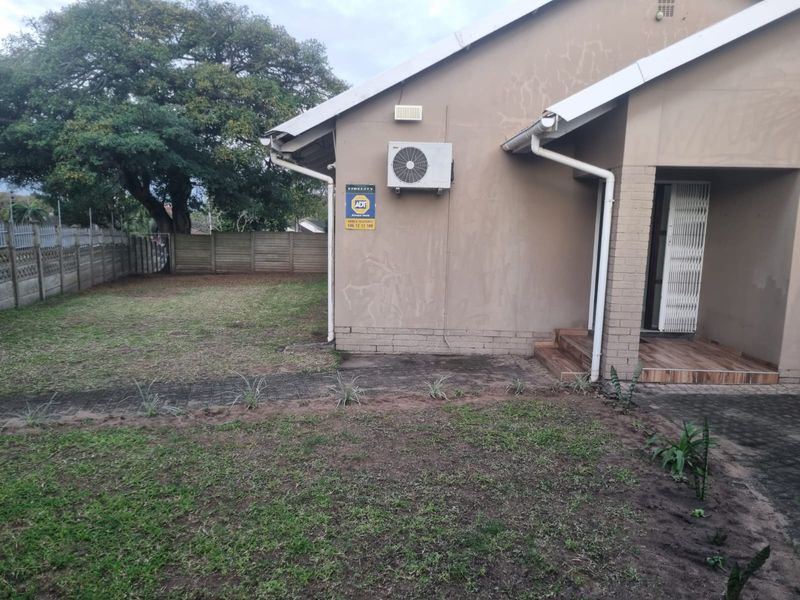 Rear exterior view of the property featuring a lawn, paved walkway, security gate, and wall-mounted air conditioning unit.
