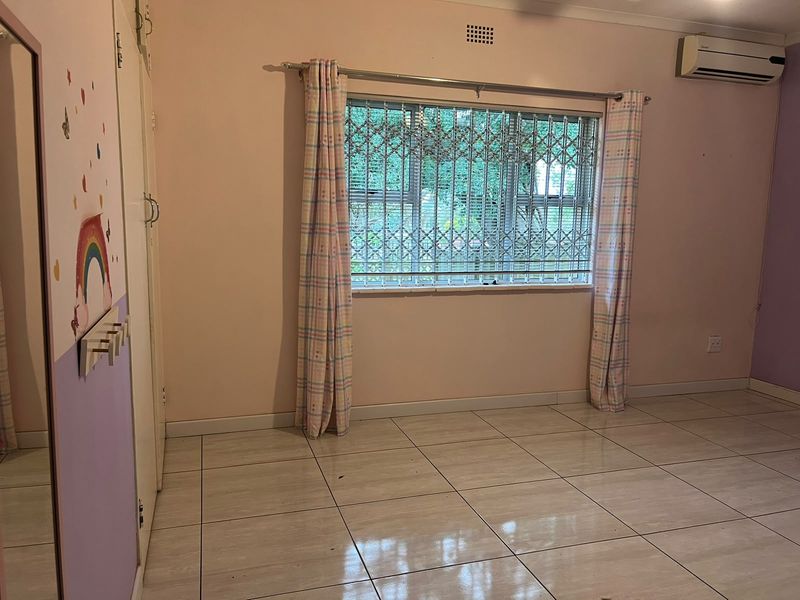 A spacious bedroom featuring tiled flooring, built-in cupboards, and an air conditioning unit, with security bars installed on the window.