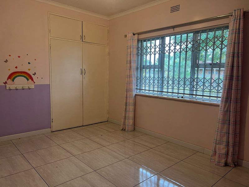 A spacious bedroom featuring tiled flooring, built-in cupboards, and a window fitted with a security gate.