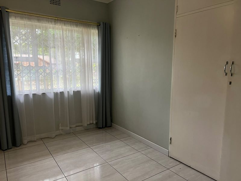 A bright, empty bedroom featuring tiled flooring, a built-in wardrobe, and a window fitted with a security gate.