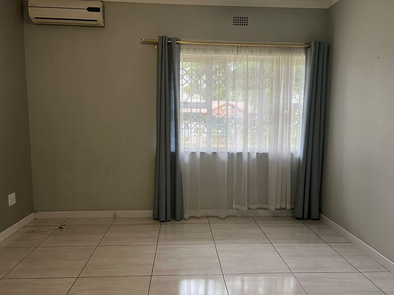 A clean, empty bedroom featuring neutral tiled flooring, a wall-mounted air conditioning unit, and a window fitted with security bars.