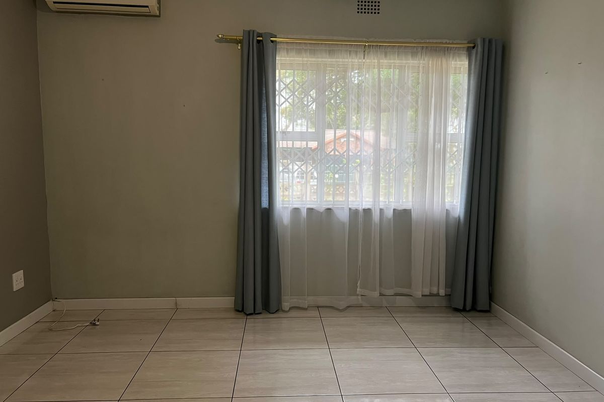 A clean, empty bedroom featuring neutral tiled flooring, a wall-mounted air conditioning unit, and a window fitted with security bars.