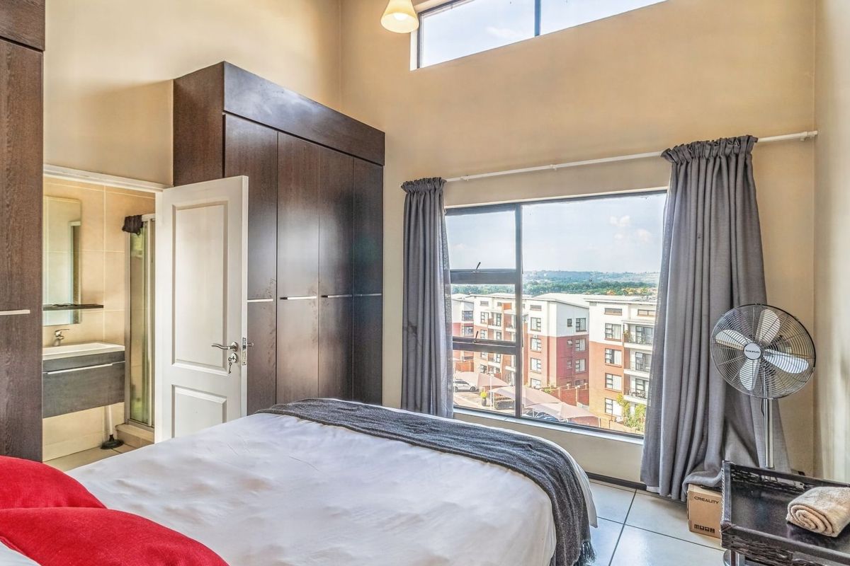 A bright bedroom featuring built-in cupboards, high ceilings with a clerestory window, and a large window offering an elevated view of the surrounding complex.