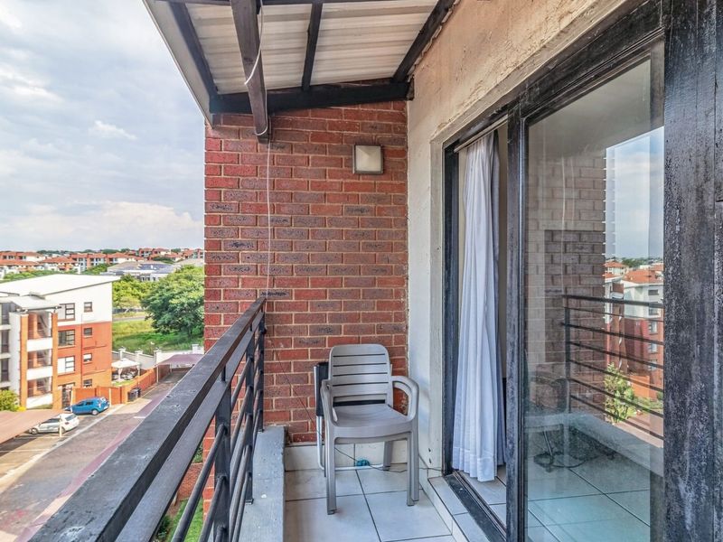 A covered balcony featuring tiled flooring, an exposed brick wall, and a metal balustrade, offering an elevated view of the surrounding apartment complex.