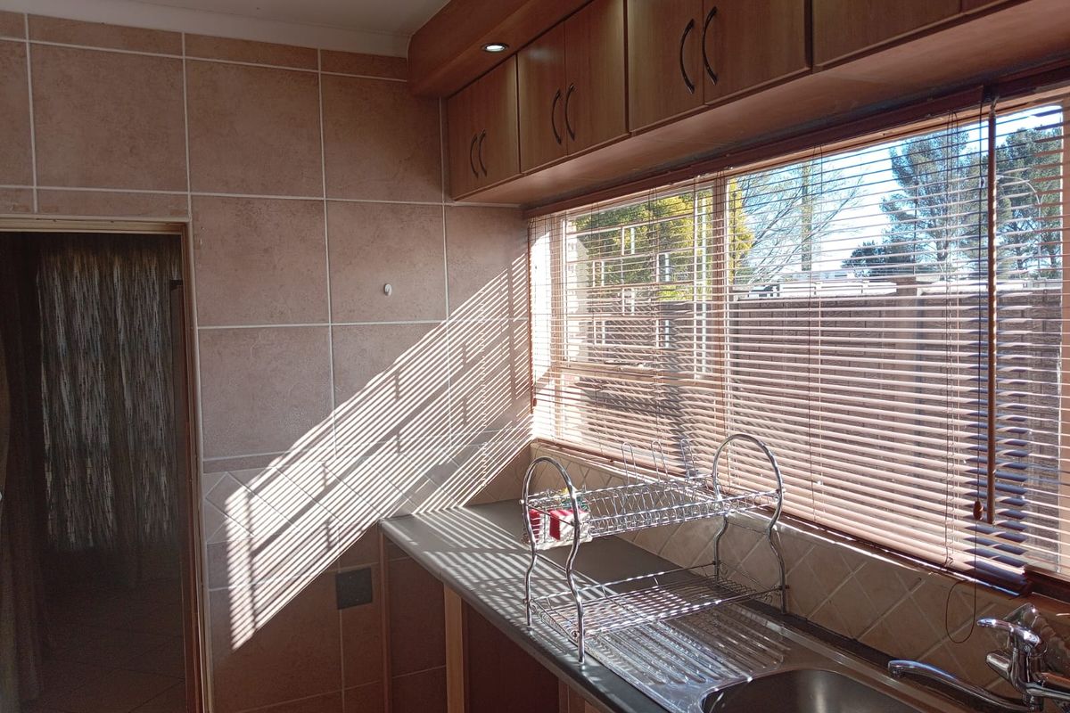 A functional kitchen area featuring wood-tone cabinetry, tiled splashbacks, a stainless steel sink, and natural light filtering through venetian blinds.