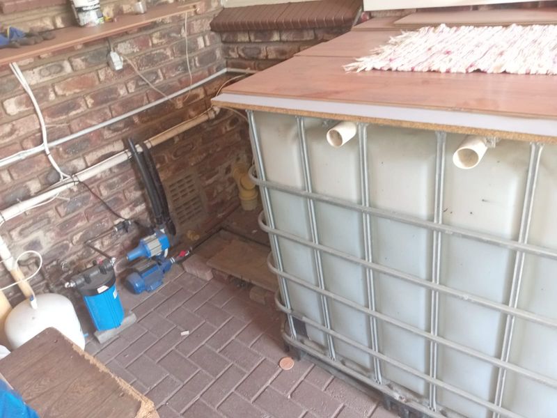Utility area featuring a water storage tank, pump, and filtration system against a brick wall with paved flooring.