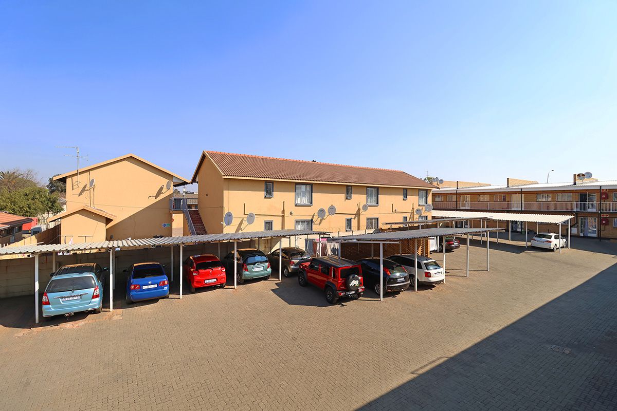 Exterior view of a townhouse complex featuring a paved parking area with covered carports and multi-story residential units.