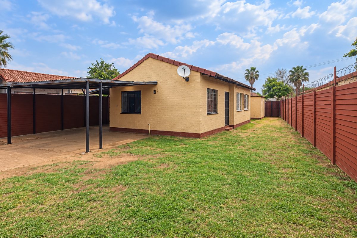Rear view of the property featuring a carport, brick boundary walls with security fencing, and a spacious garden area.
